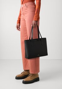 Mustat suorakaiteen muotoiset matkakassit, jotka on valmistettu teksturoidusta nahasta, joissa on kaksi kantohihnaa ja hillittyjä metalliosia, kuljetaan vaaleanpunaisien leveälahkeisten housujen rinnalla.