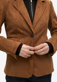 Blazer de ante marrón con cierre de un solo botón, solapas con muesca y dobladillo curvo. Superpuesto a una camiseta negra de canalé con cremallera.