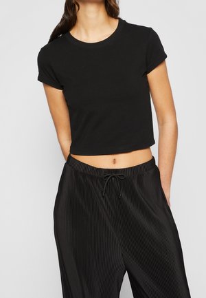 Kobieta nosząca prosty czarny krótki crop top z krótkim rękawem oraz czarne plisowane spodnie z troczkami i pionowymi paskami.