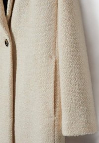 Abrigo de lana texturizado beige con un solo botón oscuro en la parte delantera, mangas largas y bolsillo lateral visible.