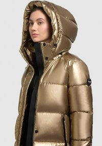 Gouden pufferjack met een glanzende, gewatteerde buitenkant, een grote capuchon en zwarte ritssluitingen en knopen. Heeft een slank, modern ontwerp.