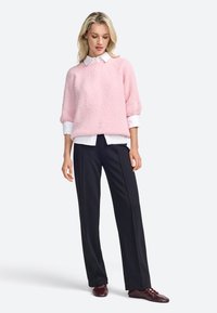 Suéter de punto rosa con un acabado texturizado, combinado con una camisa blanca de cuello y pantalones negros. El modelo lleva zapatos marrones de cordones.