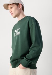 Zelená mikina s ribovanými manžetami a výstrihom, s bielym logom Calvin Klein na prednej strane. Model ju nosí s nohavicami v neutrálnych farbách.