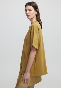 T-shirt oversize jaune moutarde à manches courtes, présentant des zips décoratifs et des accents ronds, en tissu en coton doux et texturé.