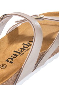 Beige lädersandal med tunna remmar, strukturerad korkfot säng och en vit gummisula. Har en metallspänne som accent. Märkesnamnet synligt.