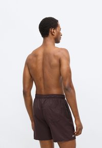 Shorts de bain marron avec une ceinture élastique, des poches latérales et arrière, et une bande d'accent verte. Texture lisse, longueur jusqu'au genou.