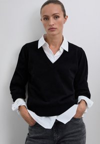 Frau mit Händen in den Taschen, die einen schwarzen V-Ausschnitt-Pullover über einem weißen Kragenhemd und grauen Jeans trägt, vor einem einfarbigen hellen Hintergrund steht.