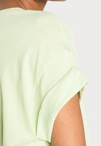 Jasnozielona koszulka o luźnym kroju, z podwiniętymi krótkimi rękawami i miękkim, teksturowanym materiałem, odsłaniająca część ramienia i górnej części ramienia.