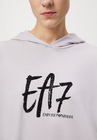 Felpa con cappuccio viola chiaro, caratterizzata da un logo "EA7" in glitter nero e dal nome del marchio "EMPORIO ARMANI" in nero sottostante. Tessuto liscio.