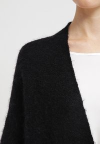 Cardigan nero lavorato a maglia in morbida lana testurizzata, con apertura frontale e silhouette drappeggiata. Design senza colletto e senza chiusure.