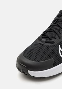 Scarpa atletica nera con tomaia in rete, superficie testurizzata, lacci neri e suola bianca. Presenta una punta arrotondata e un accento con logo.