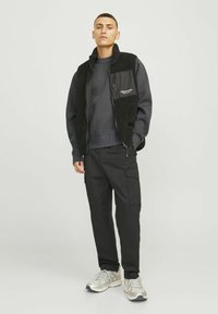 Sort fleece vest over grå sweatshirt, parret med sorte cargo bukser og sølv sneakers. Glatte teksturer med kontrasterende materialer.
