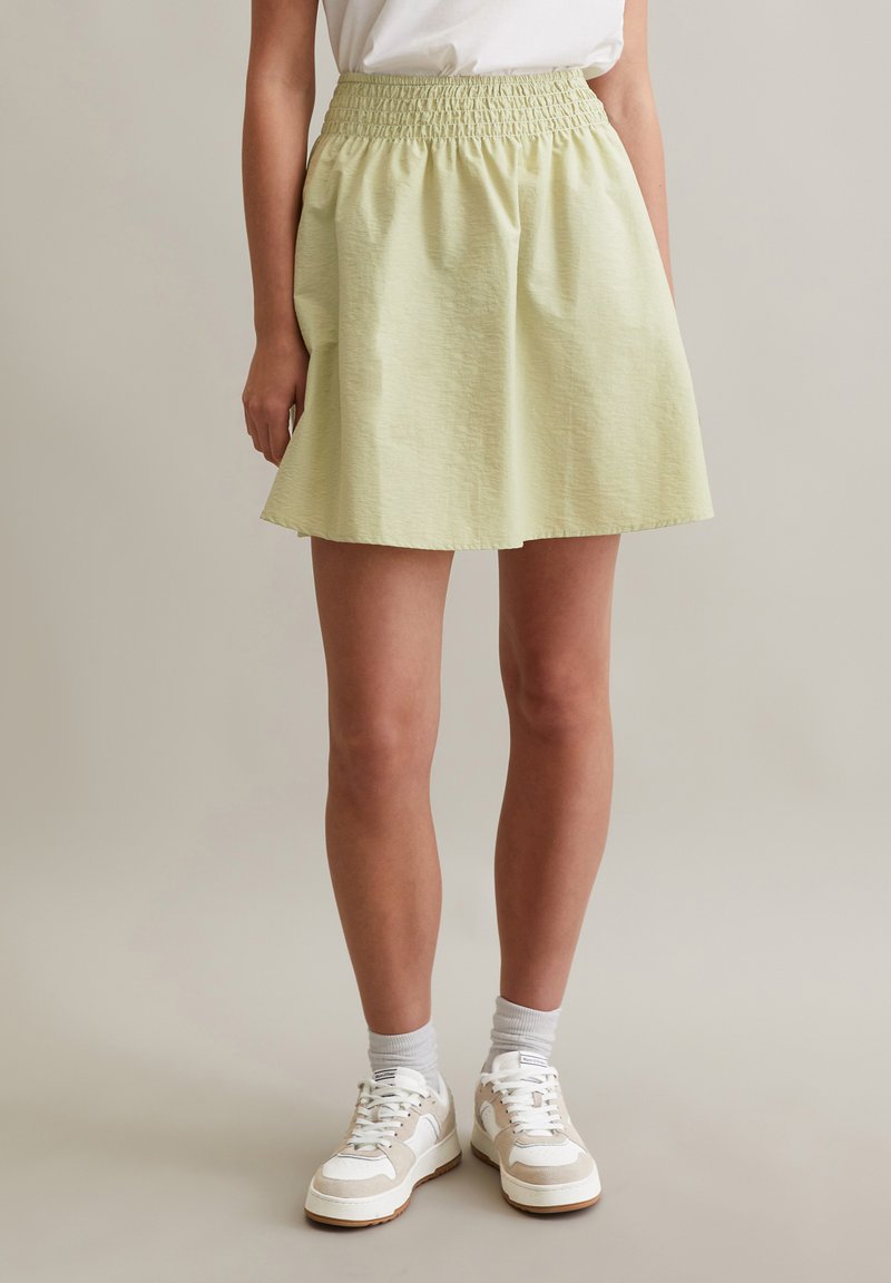 Lichtgroene, knielange rok met een smocktailleband, uitgevoerd in een gestructureerde stof en met een uitlopende vorm. Draagt comfortabel met witte sneakers.