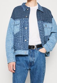 Kurtka dżinsowa w różnych odcieniach niebieskiego, z graficznymi wzorami logo, kołnierzem, przednimi kieszeniami i zapięciem na zatrzaski.