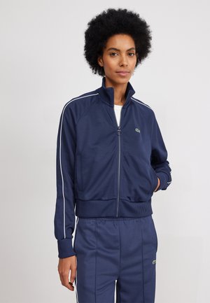Navy zip-up jacket made of smooth fabric, featuring a high collar, white side stripes, and a green logo on the left chest.