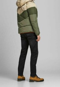 Puchowa kurtka w beżowo-zielonym wzorze kolorystycznym. Model łączy ją z czarnymi dżinsami i brązowymi butami turystycznymi.