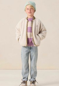 Kind dat een mintgroene beanie, een crèmekleurige fleecejas, een paars en crèmekleurig gestreept sweater, lichtere spijkerbroeken en beige sneakers draagt, met zijn handen in de zakken.