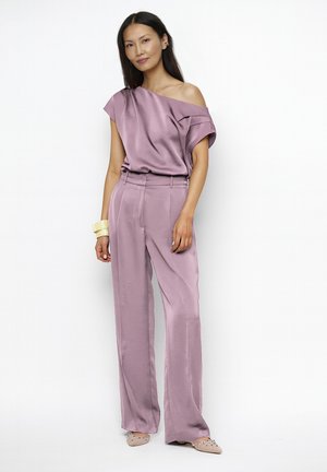 Vrouw draagt een mauve satijnen top met één schouder en bijpassende hoge taille wijde broek, geaccentueerd met crèmekleurige armbanden en nude ballerina's met versieringen.