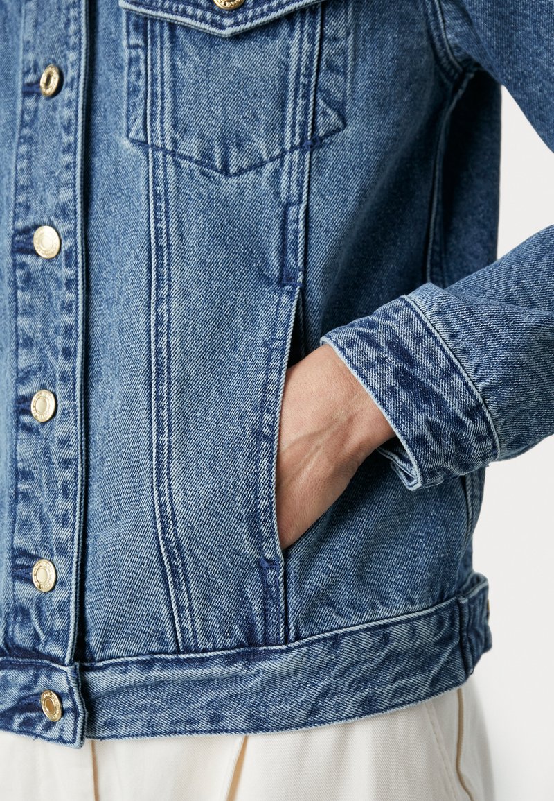 Chaqueta de mezclilla color azul claro con cuello, bolsillos delanteros y botones dorados. Presenta costuras visibles y un ajuste relajado.