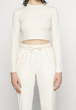 Person iført en cremefarvet langærmet crop top og matchende cremefarvede snøre bukser, stående mod en ensfarvet hvid baggrund.