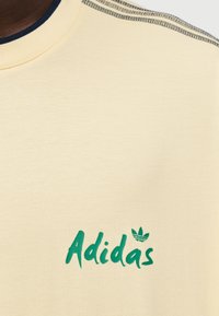 Κίτρινη βαμβακερή μπλούζα με πράσινο λογότυπο "Adidas" και τριφύλλι, με ριμπ λαιμόκοψη σε σκούρο μπλε και τόνους στους ώμους. Μαλακή, λεία υφή.