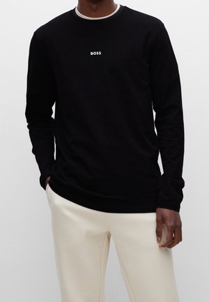 Czarna koszula z długim rękawem z białym logo „BOSS” na piersi. Okrągły dekolt z wewnętrznym białym kołnierzykiem. Połączona z kremowymi spodniami.