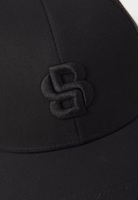 Svart caps laget av teksturert stoff har en brodert logo på framsiden. Designet inkluderer en buet skjerm og sømdetaljer.