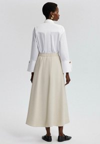 Witte blouse met lange mouwen en knoopmanchetten, gecombineerd met een beige, knie-lange rok met elastieke taille. Zwarte platte schoenen maken de look compleet.