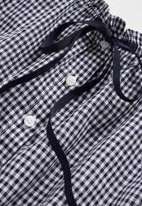 Skjorte i marineblått og hvitt rutet stoff med rynket halslinning, bånd og tre hvite knapper foran. Mykt, teksturert materiale.