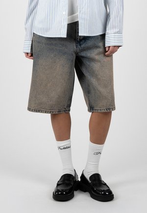 Person trägt ausgewaschene kniehohe Denim-Shorts, weiße Socken mit Schriftzug, schwarze Loafers und ein weißes gestreiftes Hemd mit langen Ärmeln.