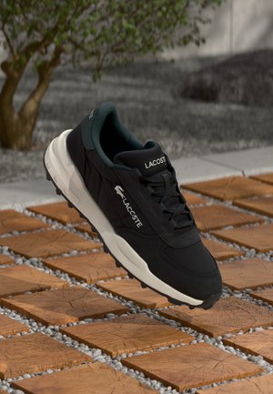 Black athletic sneaker with a textured fabric upper, contrasting dark green lining, white midsole, and Lacoste branding on the side.