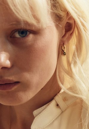 Zelta riņķa auskarā ar mazu apaļu piekariņu, kas karājas no viegli iedegusas auss. Tuva bilde ar bālas ādas un blondiem matiem seju.