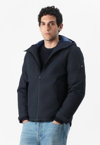 Navy hooded jacket made of sturdy material with zip closure, featuring side pockets and a blue inner lining. Accented with logo patch.