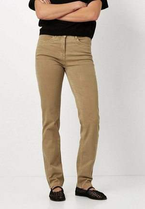 Beige, slim-fit jeans with a classic five-pocket design, button and zipper closure, and a smooth texture. Worn with black flat sandals.