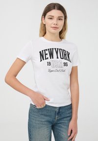 T-shirt in cotone bianco con una grafica nera audace che recita "NEW YORK 1995" con "Upper East Side" sottostante. Maniche corte e vestibilità classica.