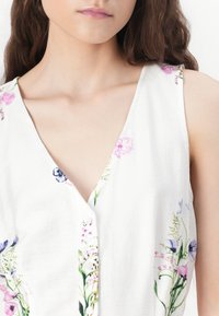 Top senza maniche in tessuto bianco con stampa floreale in rosa, blu e verde. Presenta un collo a V e dettagli con bottoni lungo la parte anteriore.