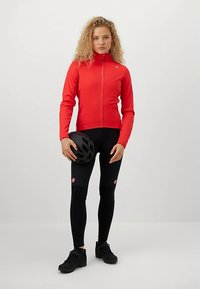 Γυναίκα που φοράει κόκκινη ζακέτα ποδηλασίας, μαύρο παντελόνι ποδηλασίας και παπούτσια, κρατώντας ένα μαύρο κράνος ποδηλασίας μπροστά σε έναν απλό ανοιχτό φόντο.