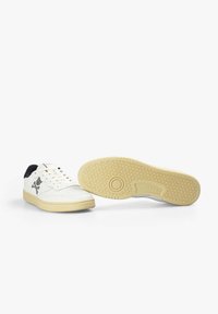 Zapatillas blancas de diseño bajo, con acentos negros en la parte trasera y una suela texturizada beige. Presenta un logotipo con una calavera y tibias.
