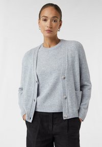 Hellgrauer Cardigan aus weichem Strickstoff, mit rundem Halsausschnitt, vorderen Knöpfen und seitlichen Taschen. Kombiniert mit einem passenden Pullover.