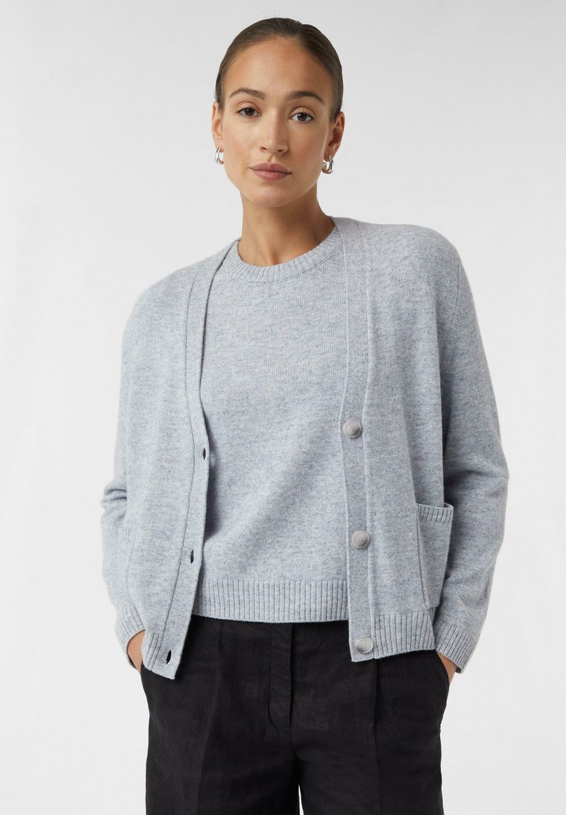 Hellgrauer Cardigan aus weichem Strickstoff, mit rundem Halsausschnitt, vorderen Knöpfen und seitlichen Taschen. Kombiniert mit einem passenden Pullover.