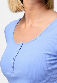 Sv ětle modrý žebrovaný top s lodičkovým výstřihem, vybavený knoflíkovou légou a krátkými rukávy. Tkanina vypadá jemně a je mírně přiléhavá.