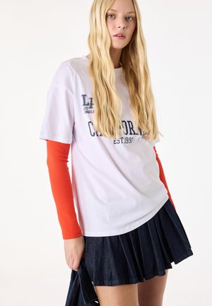 Jeune femme aux longs cheveux blonds portant un T-shirt blanc à motif California par-dessus un pull rouge à manches longues et une jupe plissée foncée.