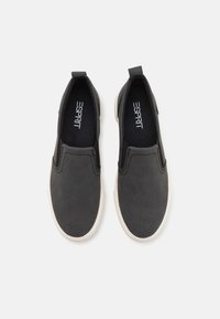 Slip-on skor tillverkade av mörkgrått texturerat material, med svarta innersulor och en vit gummisula. Har minimal sömnad och två dragflikar.