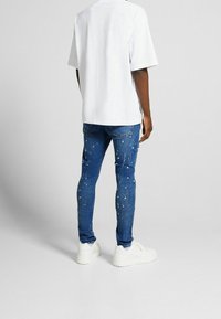 Vit bomullströja med korta ärmar, tillsammans med blå denimjeans med färgsplatterdetaljer. Vita sneakers fulländar outfiten.
