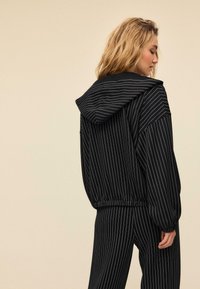 Černá mikina s kapucí a pruhy s volnějším střihem, která má kapuci a elastické manžety. Materiál vypadá měkce a má vertikální bílé tenké pruhy.