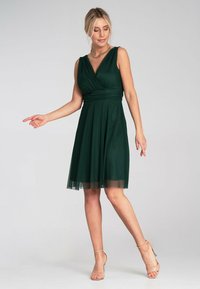 Grünes knielanges Kleid mit angepasstem Oberteil und ausgestelltem Rock. Hat einen V-Ausschnitt, Kurzärmel und einen durchsichtigen Überzug. Tanfarbene High-Heels.