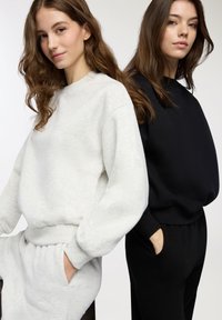 Due donne sono in piedi schiena a schiena, indossando felpe e pantaloni casual coordinati, una di colore grigio chiaro e l'altra di colore nero, con le mani nelle tasche.
