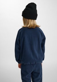 Marinblå fleece-tröja med avslappnad passform och ribbade muddar, ihopparad med matchande byxor, sedd bakifrån, toppad med en svart mössa.