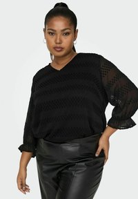 Blusa nera con scollo a V, tessuto trasparente, righe testurizzate e maniche a volant. Abbinata a pantaloni in pelle nera.