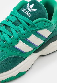 Grön sportskor med en texturerad mocka- och mesh-överdel, prydd med vita och lila ränder, rund tå och snörning.